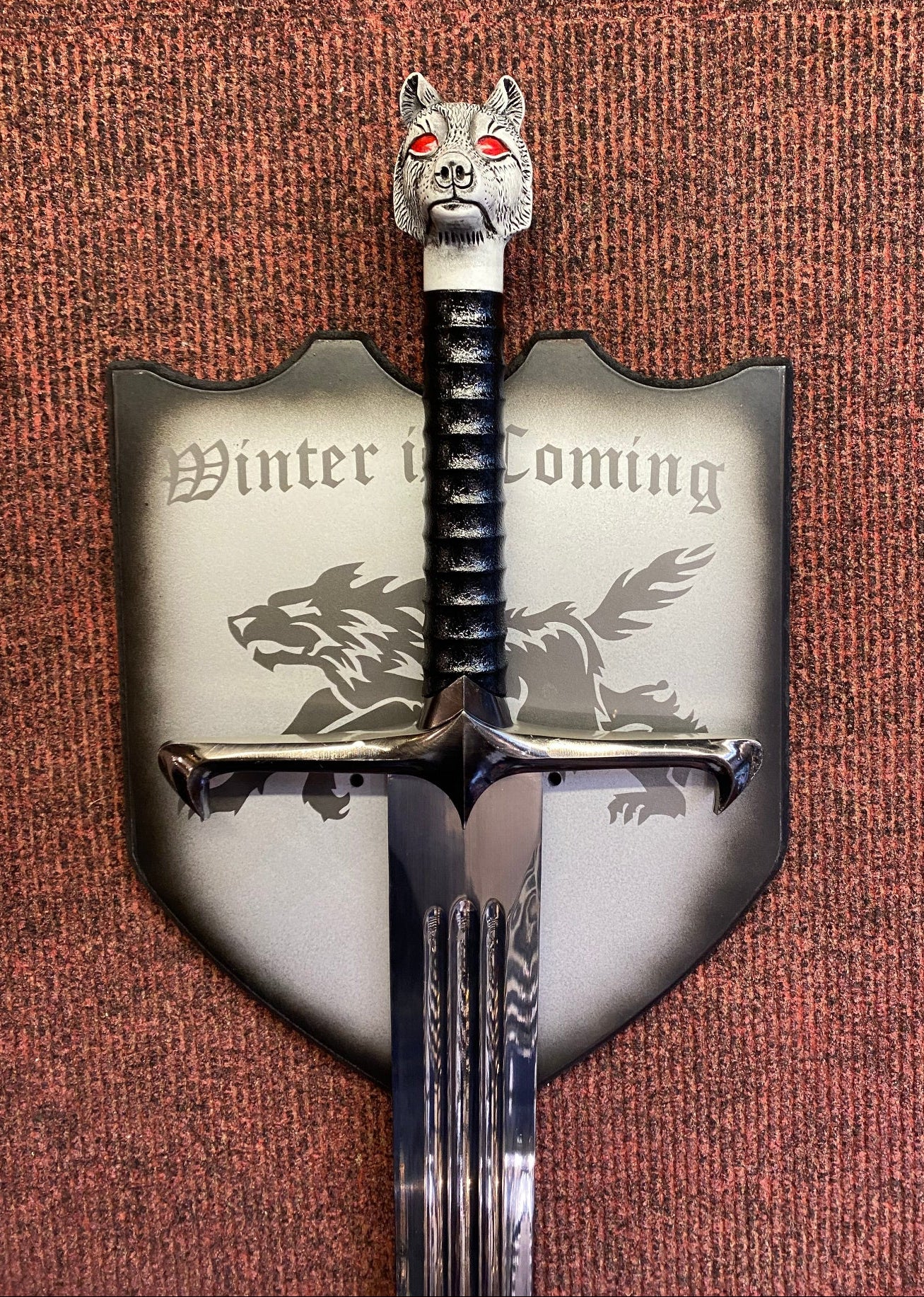 Decorative sword with a wolf head finial and 'Winter is Coming' text on a shield, against a textured brown background.