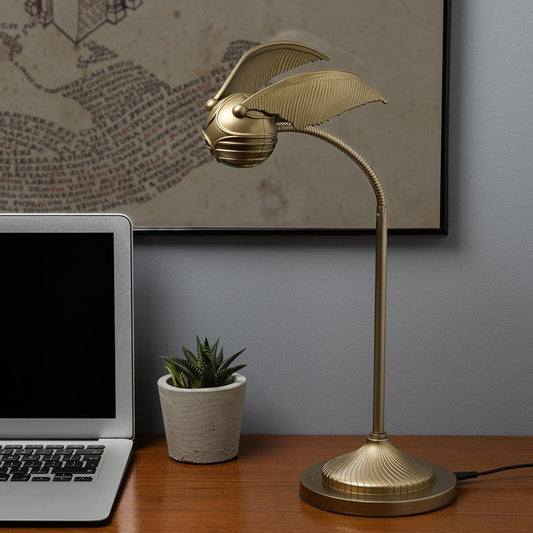 Gold desk lamp on a wooden surface next to a laptop and small plant, with a framed picture in the background.