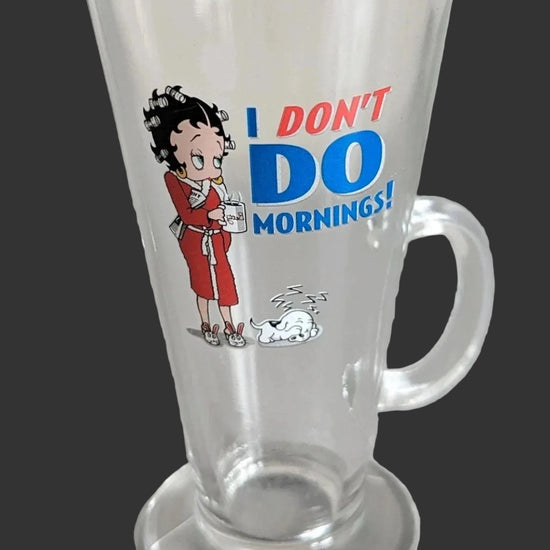 A clear glass mug with a handle and base, featuring a cartoon Betty Boop illustration and the text 'I DON'T DO MORNINGS!' in blue and red on a white studio background.