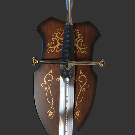 A decorative medieval-style sword with a polished silver blade, black wrapped hilt, and ornate gold detailing, shown alongside a matching dark brown wooden scabbard decorated with gold floral patterns.
