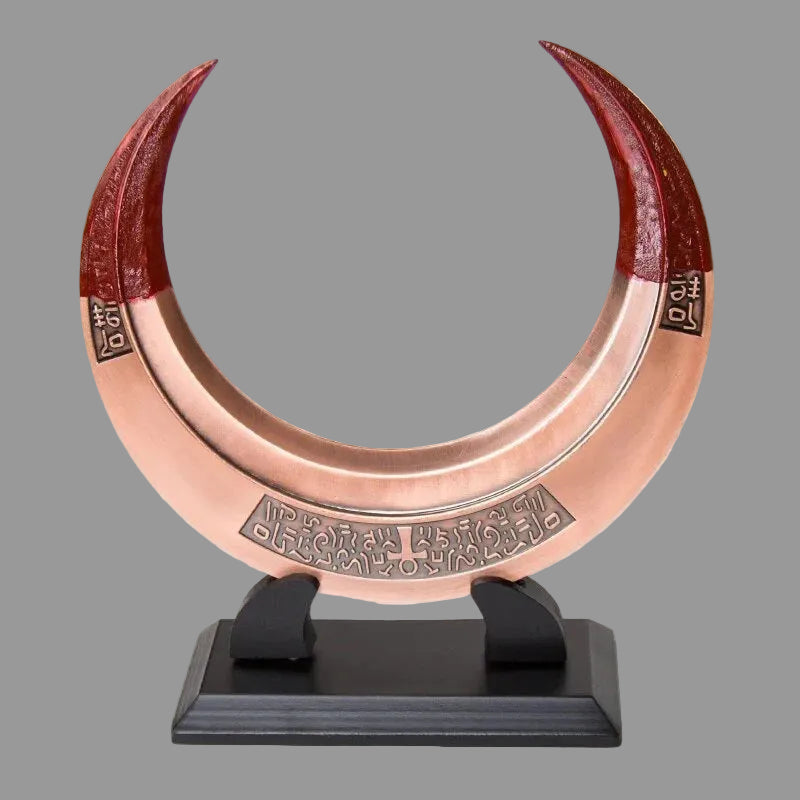 A large crescent moon pendant or wall hanging made of polished copper-toned metal with reddish-brown tips and engraved geometric details, displayed on a black stand against a plain light background.