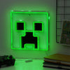 Green neon sign shaped like a character from a video game, on a wall with a controller and books below.